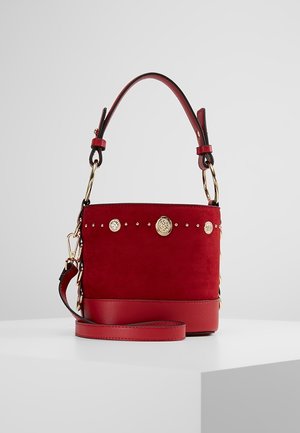 Sac seau en daim rouge avec clous et anneaux dorés, base en cuir et bandoulière ajustable, présenté sur une surface blanche avec un fond gris uni.