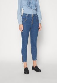 Modré denimové džíny se slim fit, s zipovým zapínáním, předními kapsami a bočními zipy na kotnících. Nosené s světle modrou blůzkou s volány.