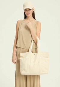 Beżowa torba na ramię z płótna z białymi akcentami i kieszenią z przodu, z napisem "Melbourne Tennis Club" w zakrzywionym wzorze.