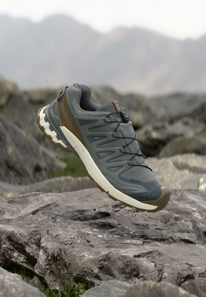 Scarpa da trail running grigia e marrone con suola robusta che fluttua sopra un terreno roccioso e uno sfondo montano sfocato.