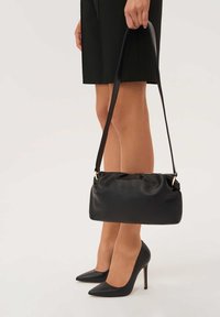 Fekete bőr kézitáska fodros tetejű, állítható pánttal és arany színű fém díszítéssel, fekete hegyes orrú magassarkúval és bézs színű lábakkal párosítva.