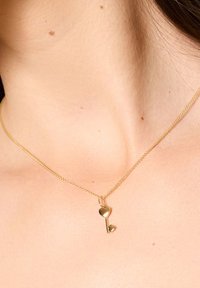 Złocisty naszyjnik z delikatnym łańcuchem, z wiszącym wisiorkiem w kształcie klucza w formie serca. Wisiorek ma polerowane wykończenie i mały akcent w postaci serca.