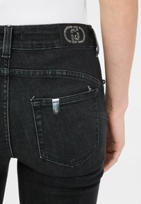 Czarne dżinsy o dopasowanym kroju, z gładką fakturą, z naszywką z logo zdobioną kryształkami oraz małym srebrnym detalem metalowym na kieszeni.