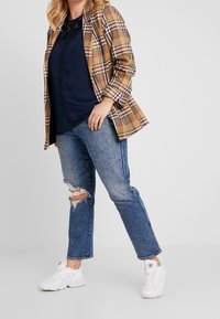 Blazer à carreaux marron et beige sur un blouse bleu marine, associé à un jean bleu déchiré et des baskets blanches, mettant en valeur les textures superposées et le style décontracté.