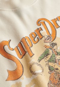 Crèmekleurige sweatshirt met een oranje "SuperDry" logo, geïllustreerd cowboymotief met groene en bruine accenten, en een geribbelde halslijn.