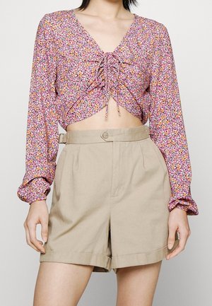 Kobieta nosząca różowy, kwiatowy crop top z długim rękawem, z wiązaniem z przodu oraz beżowe, wysokie szorty z pętlami na pasek i zapięciem na guzik.