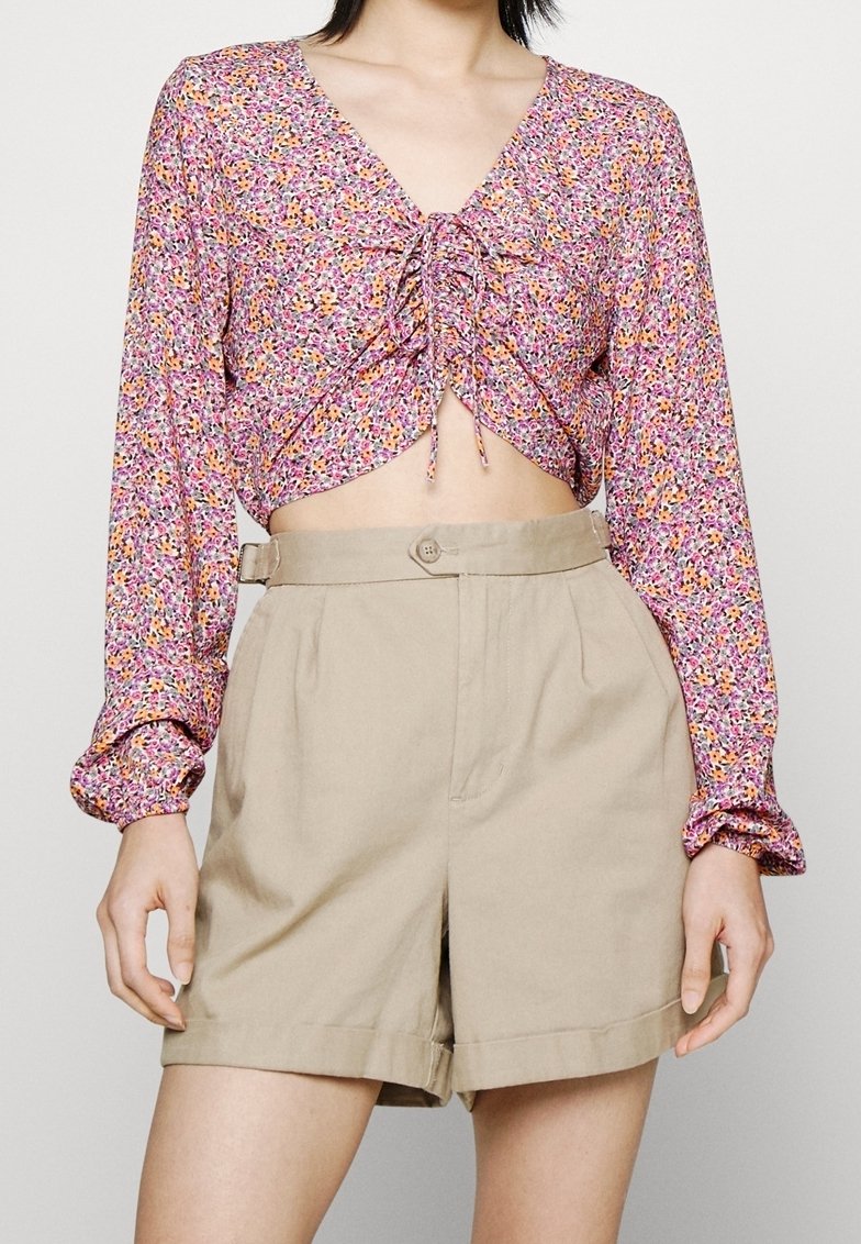 Femme portant un crop top fleuri rose à manches longues avec un nœud à l'avant et un short beige taille haute avec passants de ceinture et fermeture par bouton.