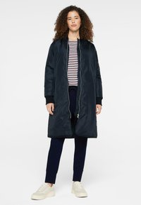 Dunkelblauer, langer wattierter Mantel mit Reißverschluss, Seitentaschen, gerippten Bündchen, getragen über einem gestreiften Hemd, kombiniert mit marineblauen Hosen und hellen Sneakern.