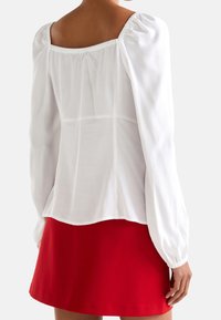 Biała bluzka z długim rękawem o kwadratowym dekolcie, z marszczonymi ramionami i przepływowym fasonem, zestawiona z czerwoną mini spódnicą.