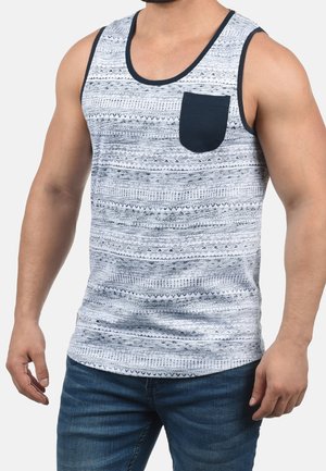 Homme portant un débardeur gris et blanc à motifs sans manches avec une poche poitrine noire et un jean bleu sur fond clair.