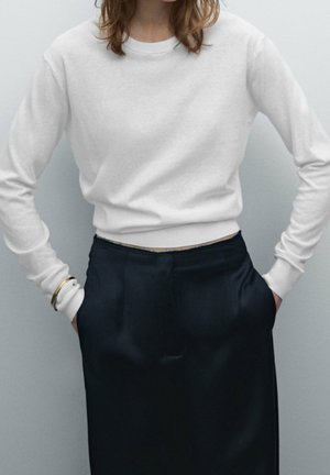 Femme portant une chemise blanche ajustée à manches longues rentrée dans un pantalon noir taille haute, mains dans les poches, sur un fond uni.