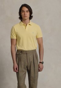 Jauns vīrietis ar plecu garuma matiem, ģērbies dzeltenā polo krekla un augsto brūno karotīšu svārku biksēm, stāv uz vienkārša fona.