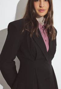 Črn tailored blazer z dvojnimi gumbi in ostrimi reverji, preplastitvijo nad svetlo vijolično prugasto srajco. Gladka tekstura z strukturiranim krojem.