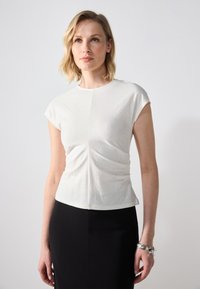 Hvit kortermet topp med en tettsittende silhuett, med en sentral søm og lett rynking ved midjen, laget av et lett stoff.