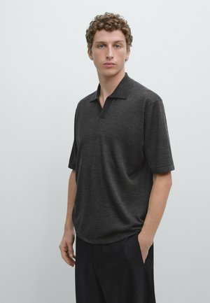 Ung mand med krøllet hår iført en mørkegrå kortærmet polotrøje og sorte bukser, stående mod en ensfarvet lys baggrund.