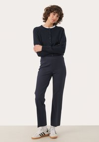 Cardigan bleu marine avec fermeture à boutons, associé à un pantalon bleu marine ajusté. Modèle portant des baskets blanches avec des rayures noires et des chaussettes blanches.