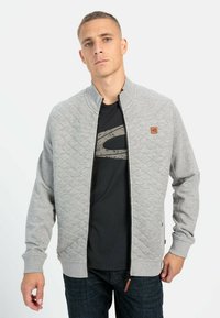 Veste matelassée grise avec un col montant, fermeture éclair à l'avant et écusson logo. Portée sur un T-shirt noir et associée à un jean foncé.