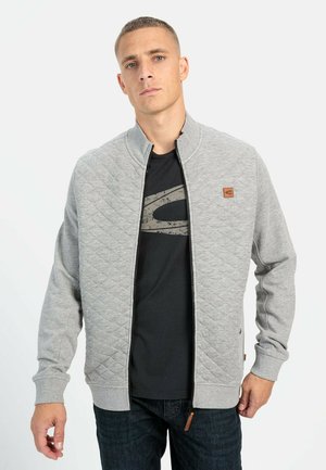 Grå quiltet jakke med høj krave, lynlås foran og logo-patch. Bæres over en sort T-shirt og kombineres med mørke jeans.