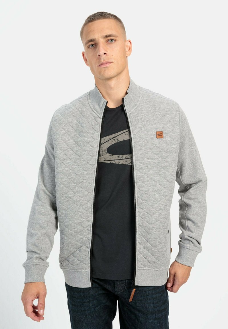 Veste matelassée grise avec un col montant, fermeture éclair à l'avant et écusson logo. Portée sur un T-shirt noir et associée à un jean foncé.