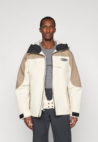 Homem de casaco exterior em blocos de cores bege e creme com colarinho preto, camisa cinza e calças cinzas escuras em fundo simples.