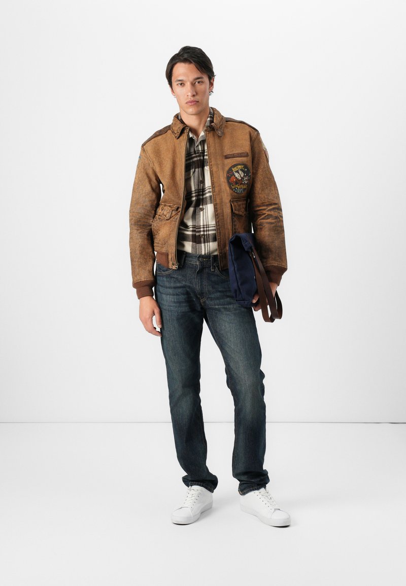 Giubbotto bomber in pelle marrone con toppa, camicia a quadri, jeans in denim scuro e sneakers bianche, che tiene una borsa a tracolla blu navy.