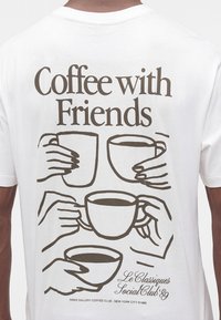 Λευκό μπλουζάκι με καφέ κείμενο "Coffee with Friends" και εικονογραφήσεις τεσσάρων χεριών που κρατούν φλιτζάνια καφέ στην πλάτη.