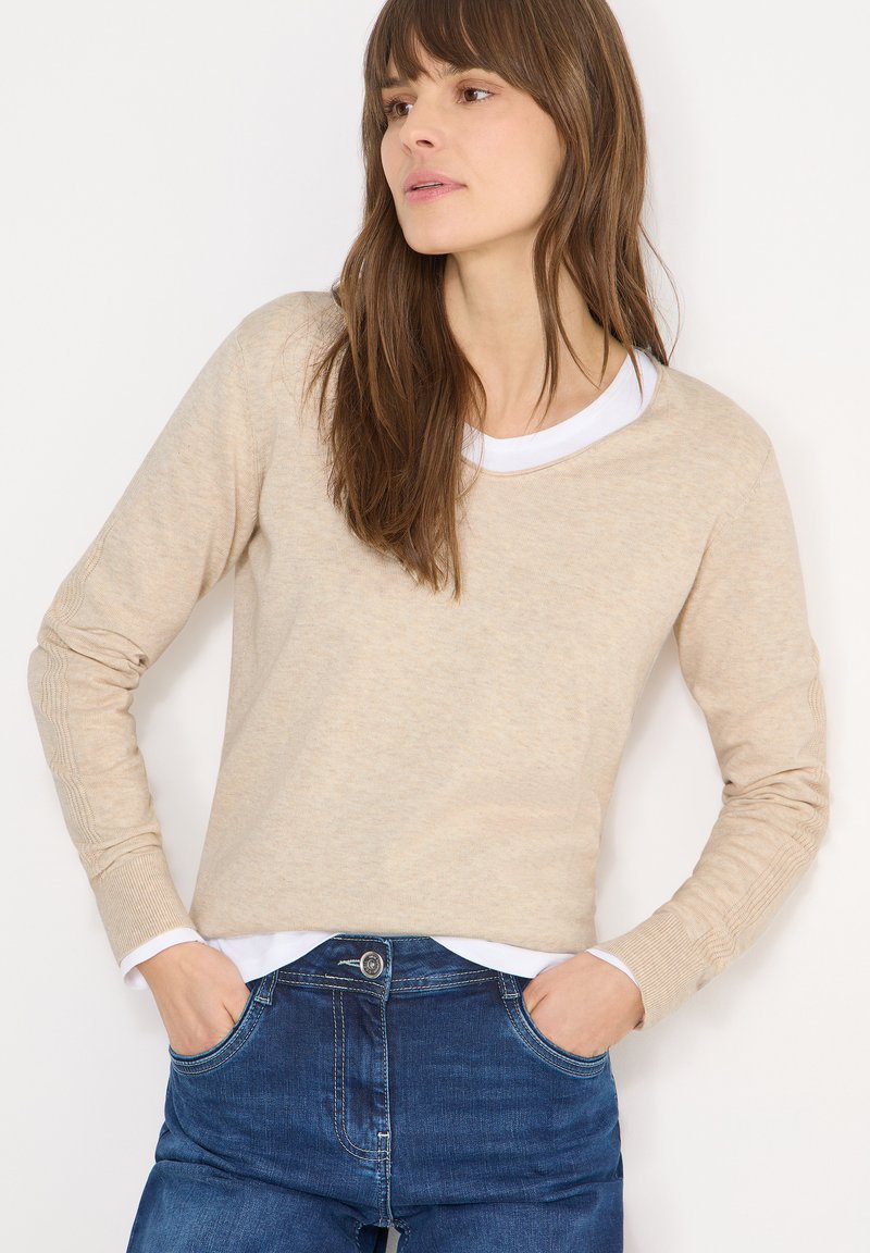 Pull tricoté beige à manches longues et col rond, superposé à un T-shirt blanc. Le tissu semble doux, avec des poignets côtelés.