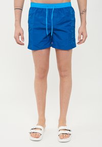 Bañador azul con cintura ajustable, logo "SUNDEK" en el lateral y tejido suave y ligero. Combinado con sandalias deslizables blancas.