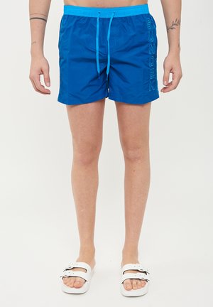 Pantaloni da bagno blu con vita regolabile, logo "SUNDEK" laterale e tessuto morbido e leggero. Abbinati a sandali bianchi.