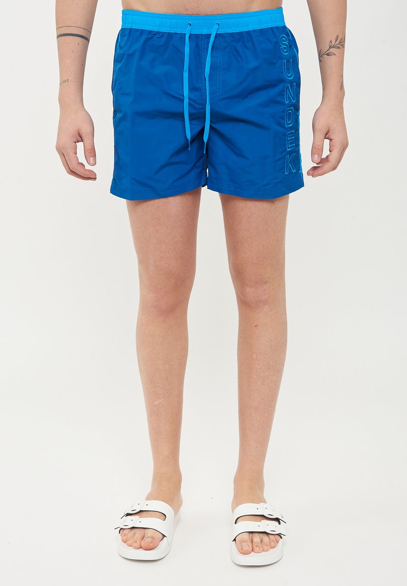 Bañador azul con cintura ajustable, logo "SUNDEK" en el lateral y tejido suave y ligero. Combinado con sandalias deslizables blancas.