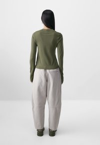 Haut à manches longues vert olive avec une silhouette ajustée et des panneaux latéraux texturés, associé à un pantalon large gris clair et des chaussures vertes.