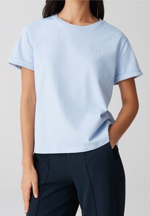 Femme portant un t-shirt bleu clair à manches courtes avec un petit emblème en forme de cœur, associé à un pantalon bleu marine foncé, main dans la poche.