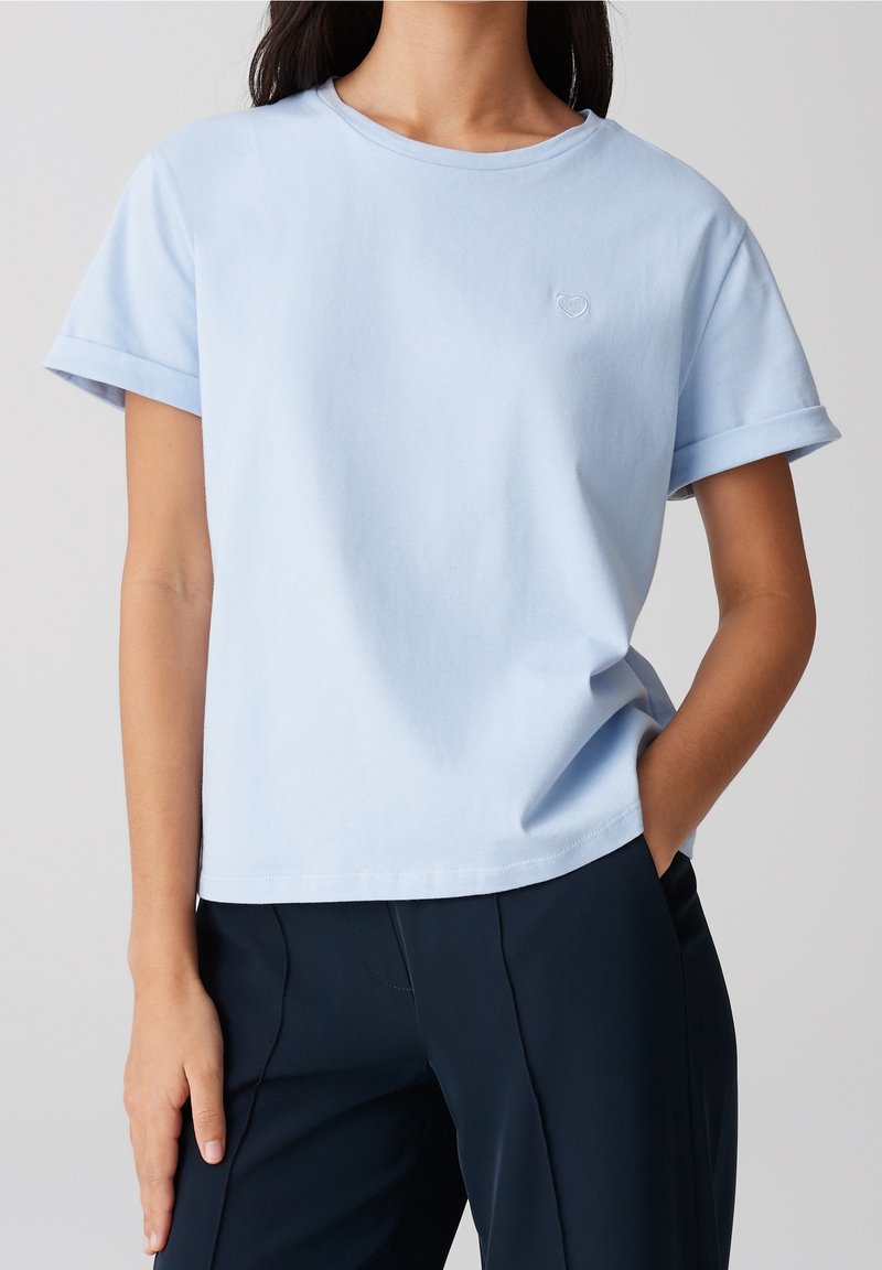 Femme portant un t-shirt bleu clair à manches courtes avec un petit emblème en forme de cœur, associé à un pantalon bleu marine foncé, main dans la poche.