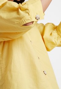 Geel overhemd met knopen, gemaakt van lichtgewicht stof. Heeft opgeblazen mouwen, ronde knopen en een ontspannen pasvorm met een geprofileerde afwerking.