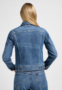 Veste en denim bleu moyen avec une coupe structurée, poignet à un bouton et deux poches arrière. Caractéristiques : col classique et détails surpiqués.
