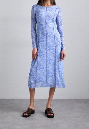 Femme portant une robe midi à manches longues, bleu clair à fleurs, et des sandales à talons blocs ouvertes à l'avant, marron foncé, debout sur un sol gris.