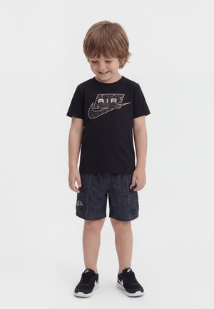 Jeune garçon portant un t-shirt noir Nike Air, un short foncé à motifs et des baskets Nike noires, souriant et regardant vers le bas.