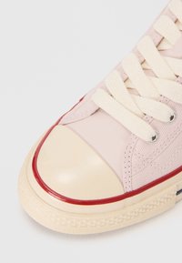 Gros plan d'une basket en toile rose avec des lacets blancs, un embout en caoutchouc beige et une bordure rouge le long du bord de la semelle.