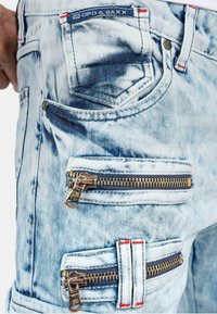 Light blue denim trousers featuring distressed texture, multiple zippered pockets, and red stitching accents. Brand label visible on waistband.