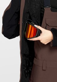 Gogle narciarskie z pomarańczowymi soczewkami, czarną ramą i regulowanym paskiem, trzymane przed kieszenią kurtki z czarnym wykończeniem i brązowymi ogrodniczkami.