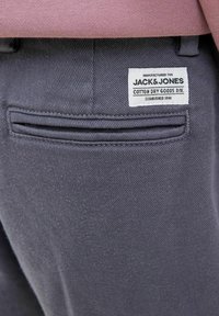 Grå bomullsshorts med en bakficka, som har en vit etikett med texten "JACK & JONES" och sömdetaljer längs fickans kant.
