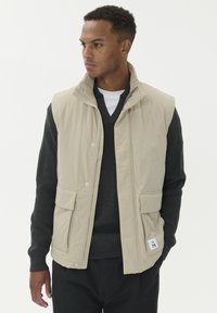 Muškarac gleda lijevo, nosi bež prsluk bez rukava preko tamnosivog V-izreza sv sweatera i crnih hlača, postavljen na jednostavnu bijelu pozadinu.