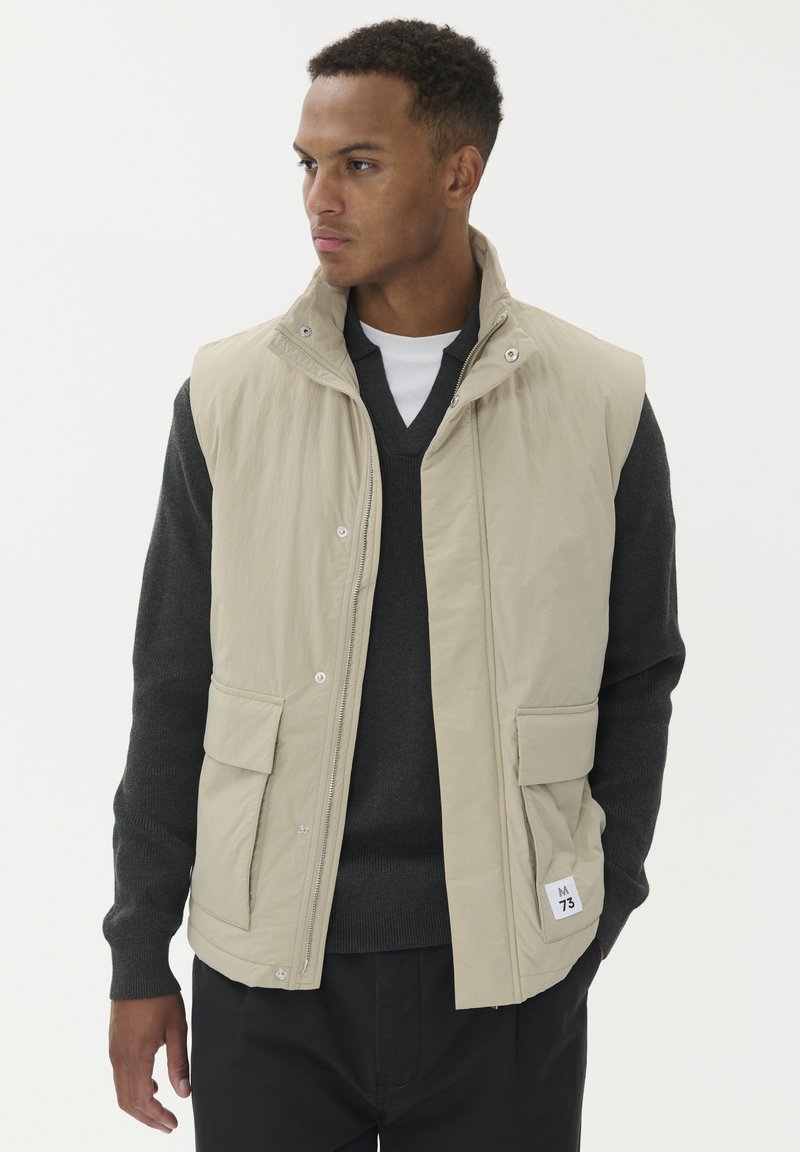 Muškarac gleda lijevo, nosi bež prsluk bez rukava preko tamnosivog V-izreza sv sweatera i crnih hlača, postavljen na jednostavnu bijelu pozadinu.