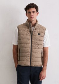 Beige gesteppte Steppweste mit Steppmuster, hohem Kragen, Reißverschluss vorne und zwei Seitentaschen; getragen über einem weißen T-Shirt, mit dunkelblauer Hose.