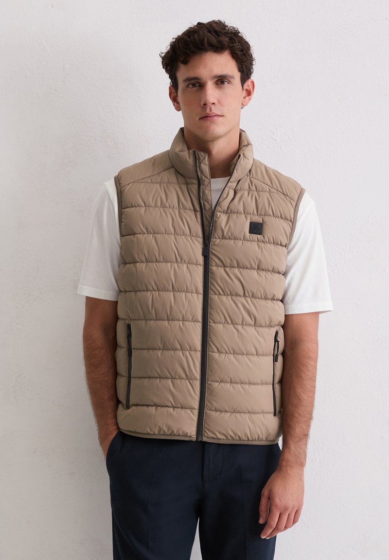 Beige gesteppte Steppweste mit Steppmuster, hohem Kragen, Reißverschluss vorne und zwei Seitentaschen; getragen über einem weißen T-Shirt, mit dunkelblauer Hose.