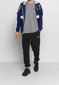 Nike větrovka v námořnické modři a bílé, spárovaná se šedým tričkem a černými tepláky. Sportovní boty dotvářejí outfit.