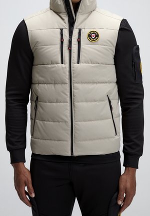 Gilet matelassé beige avec un col montant, des poches zippées et des accents noirs. Présente un patch logo sur le côté gauche de la poitrine et du bras.
