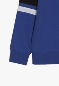 Blaues Sweatshirtärmeln mit geripptem Bündchen, das in der Nähe der Schulter schwarz-weiße Streifen zeigt, auf einem schlichten weißen Hintergrund.