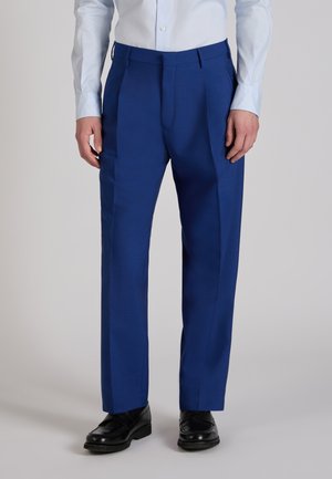 Uomo che indossa pantaloni formali blu acceso, camicia bianca elegante e scarpe di pelle nere, in piedi su uno sfondo grigio uniforme.