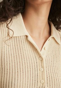 Polo shirt in maglia beige con un motivo testurizzato, colletto corto e chiusure a cinque bottoni. Il tessuto è leggero e traspirante.